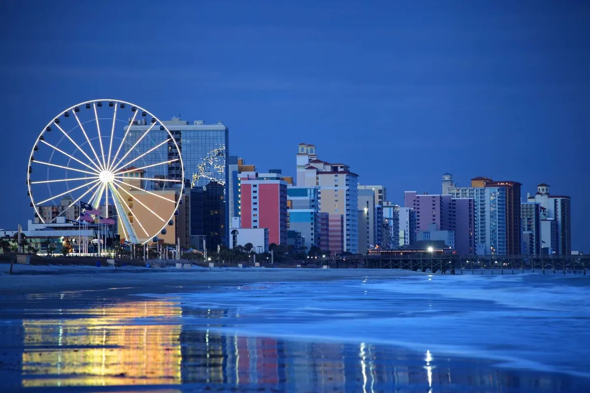 A festive beachfront scene featuring a Ferris wheel, colorful buildings, and reflective ocean waters. Families enjoy amusement rides and beach walks, with opportunities for dining and sunset views nearby. The lively, playful mood invites exploration.