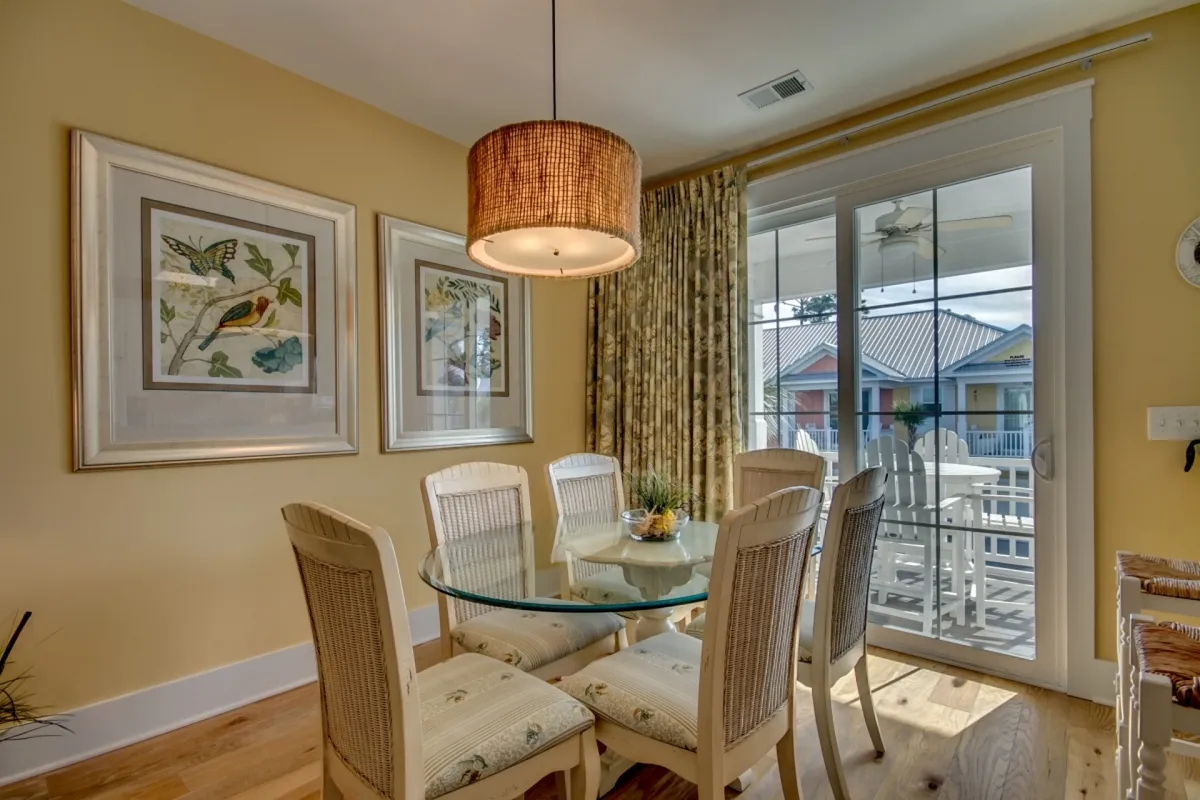 A sunlit dining area with a glass-top table, woven pendant light, and framed artwork. The area is adorned with soft curtains and offers a view of the outdoors, emphasizing the space’s warmth and adaptability for family meals or morning coffee.