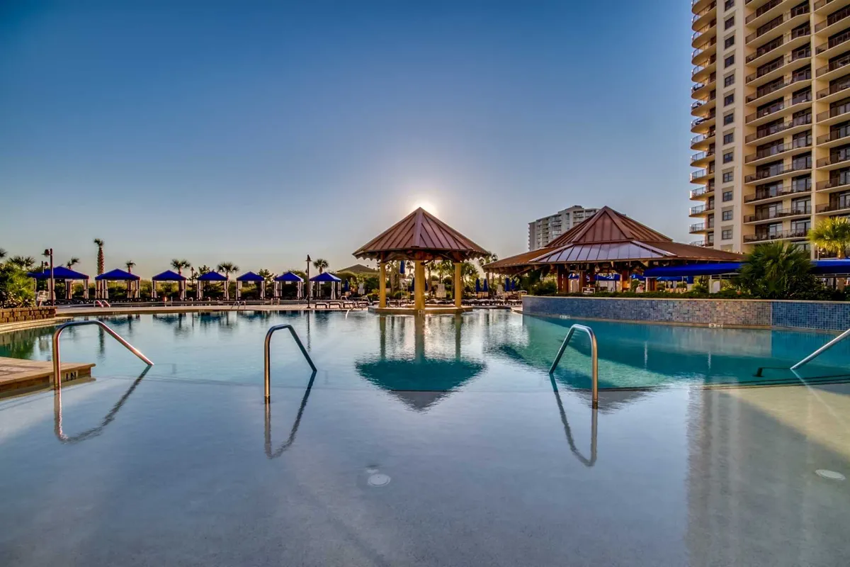 A vibrant outdoor pool area with sparkling water, thatched-roof cabanas, tall palm trees, and a high-rise resort building as a backdrop. The scene is set during sunset, enhancing the tranquil, resort-style atmosphere ideal for relaxation after active pursuits.