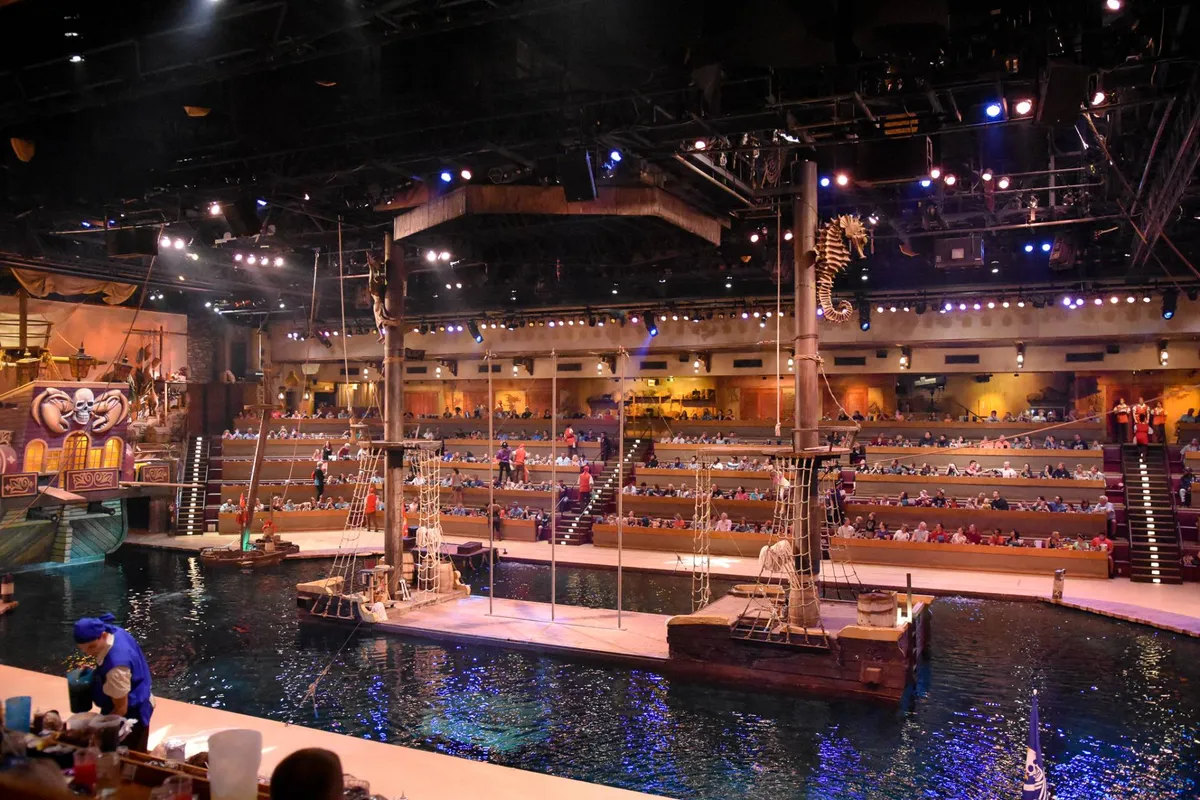 An interior view of a large audience gathered before a stage featuring water effects and a decorative ship, suggesting an immersive dinner show or live performance experience enjoyed by families and groups.