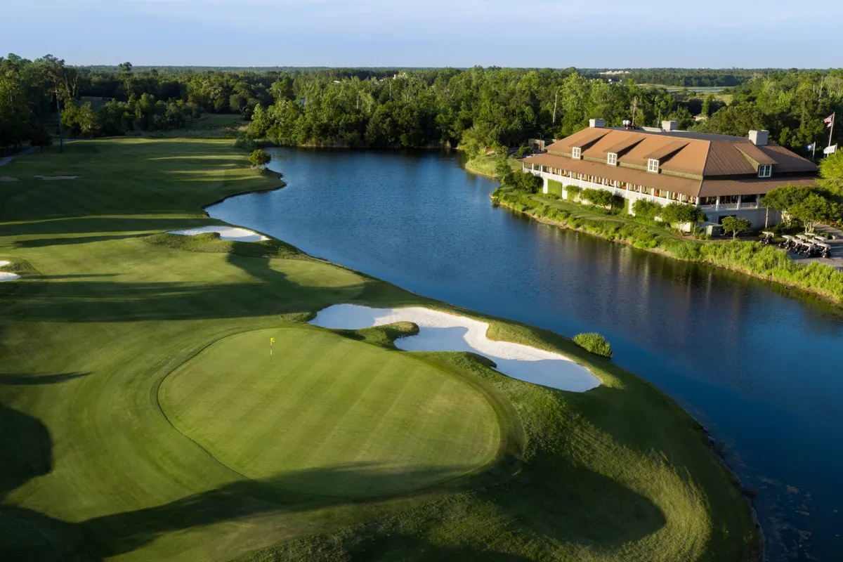 A panoramic exterior view of a lush golf course bordered by a shimmering water body. In the distance, a welcoming clubhouse is visible, hinting at community events and post-game socializing. The setting is tranquil, with manicured greens and gentle sunlight suggesting ideal conditions for both morning and afternoon play.