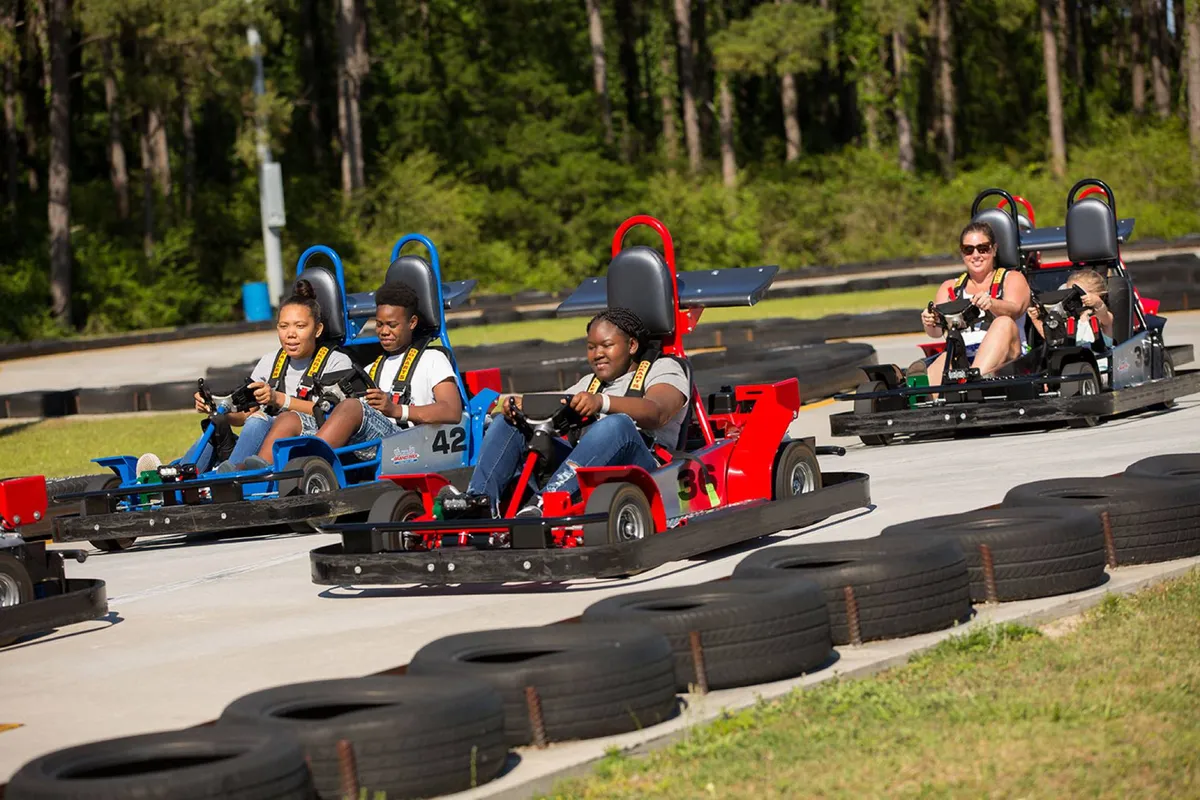 The image captures a lively go-kart racing track surrounded by greenery, with families and children participating in friendly competition. The outdoor scene exudes excitement and playfulness, spotlighting family bonding and thrilling adventures.