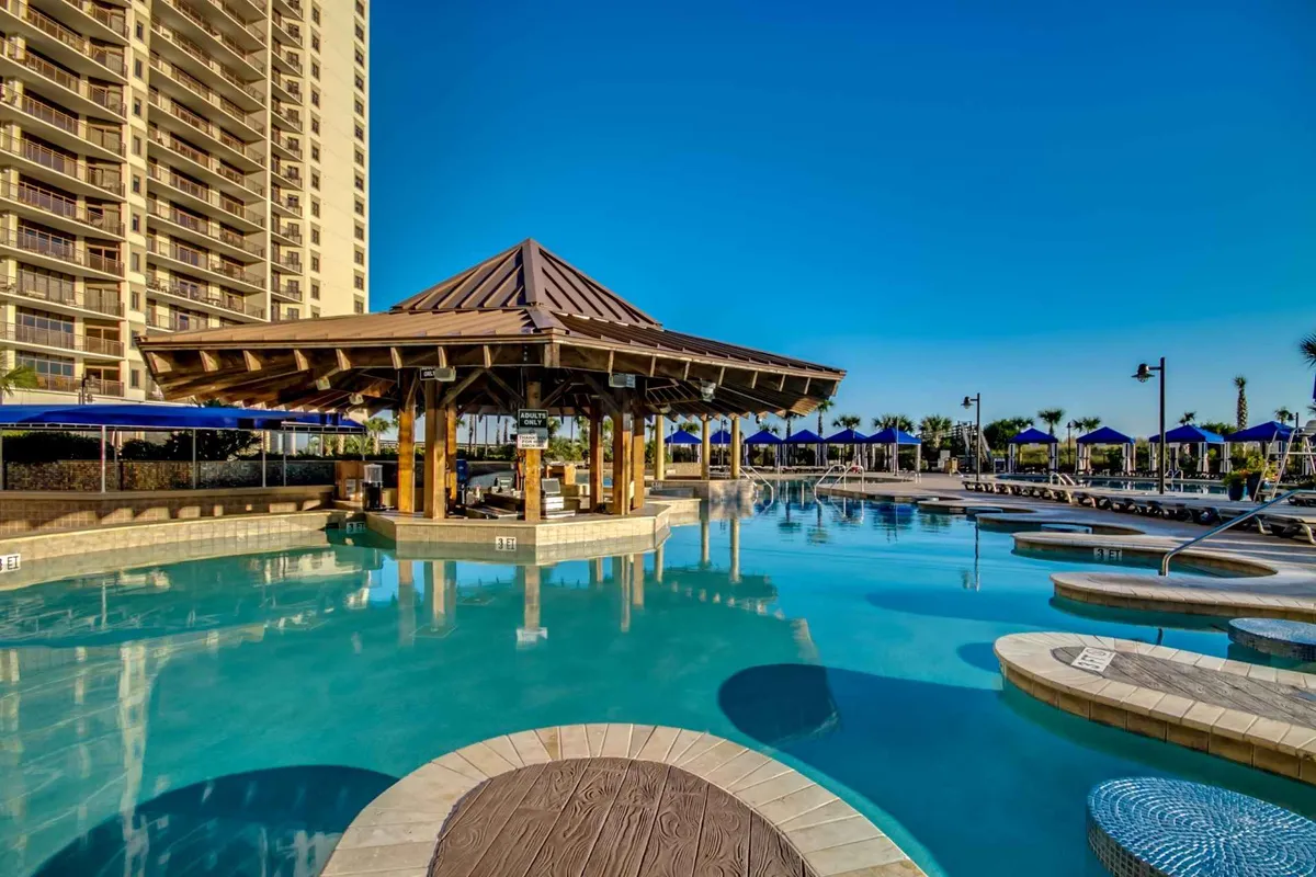 This exterior image showcases a sprawling pool complex complete with a pool, cabana, and plentiful lounge chairs under a broad sky. The scene conveys sun-soaked relaxation as families swim, sunbathe, and socialize, with the inviting cabana and hint of a swim-up bar creating a resort atmosphere.