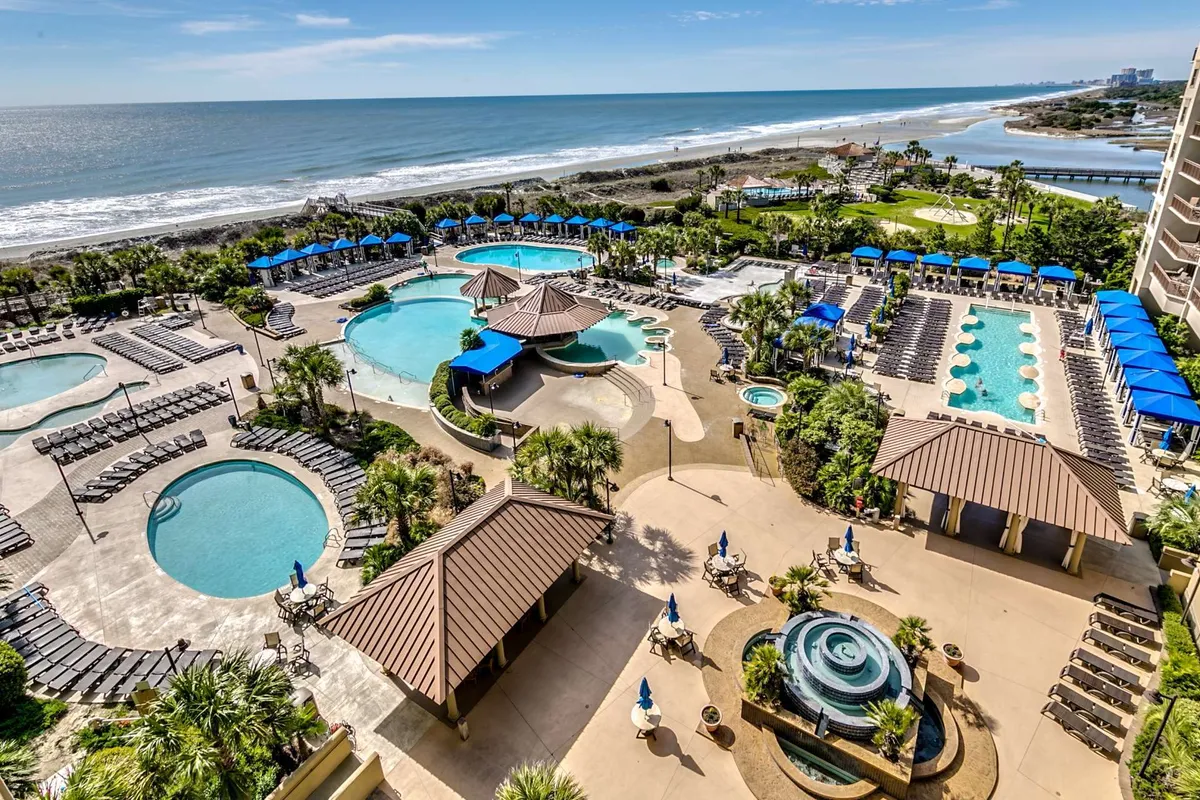 An inviting exterior scene highlighting multiple swimming pools, a hot tub, palm trees, and a beachfront view. The area is adorned with lounge chairs, inviting guests to relax in the sun or shade. The second-floor vantage is implied by the perspective over the pool area, reinforcing the sense of elevation and resort ambiance.