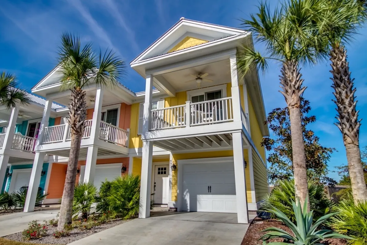 The image showcases a cheerful yellow condo with crisp white trim, framed by swaying palm trees. A spacious balcony invites guests to relax and take in the tranquil surroundings, perfect for enjoying sunsets or spending time outdoors with friends and family. The warm, coastal setting hints at leisurely days and memorable gatherings.
