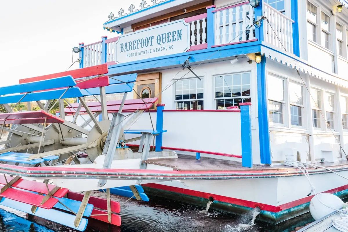 This exterior image features a colorful riverboat with a prominent paddle wheel, lively signage, and outdoor dining areas set against a scenic waterfront backdrop. The vibrant colors and bustling atmosphere evoke the excitement of riverside dining and entertainment, reminiscent of the lively ambiance found at Barefoot Landing and similar local hotspots.